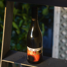 Load image into Gallery viewer, A wine bottle with a colorful orange and gold label resting on the step of a ladder outside
